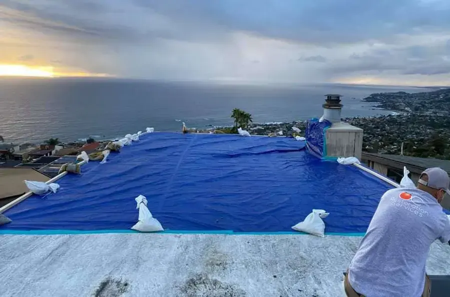 Worker securing a blue emergency tarp with sandbags on a flat roof above the ocean after a winter storm